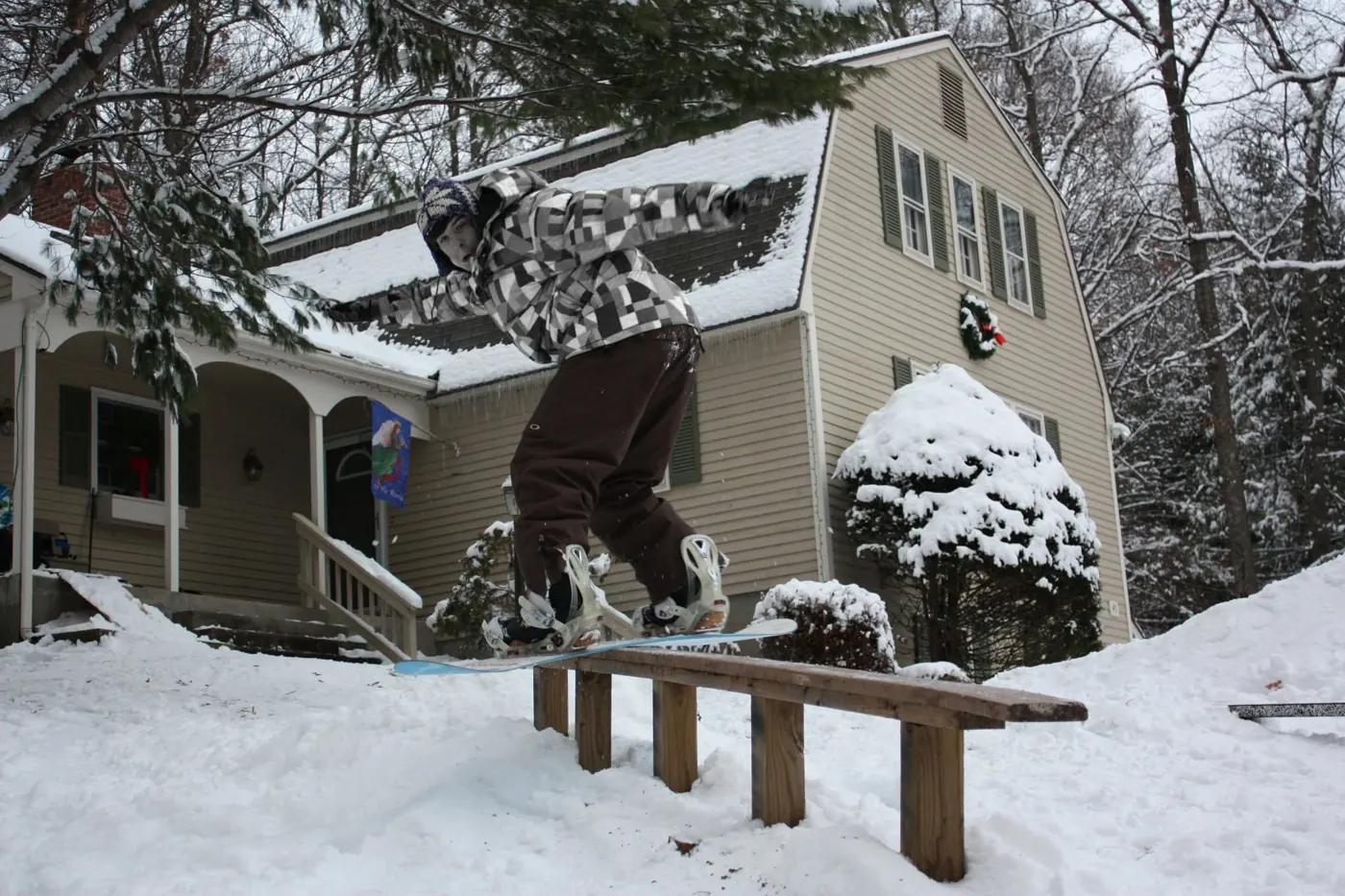 A snowboarder sliding a wooden rail in front of a snow-covered house.