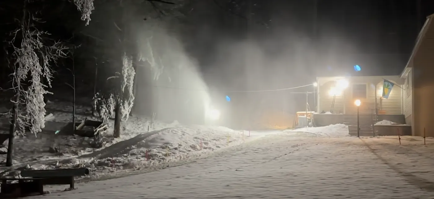 A snowgun running at night in a back yard, throwing a plume of snow under a light.